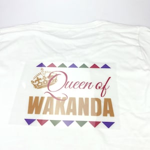 May include: A white t-shirt with a gold and red iron-on design that says "Queen of Wakanda" with a gold crown above the text. The design is surrounded by a border of colorful triangles.