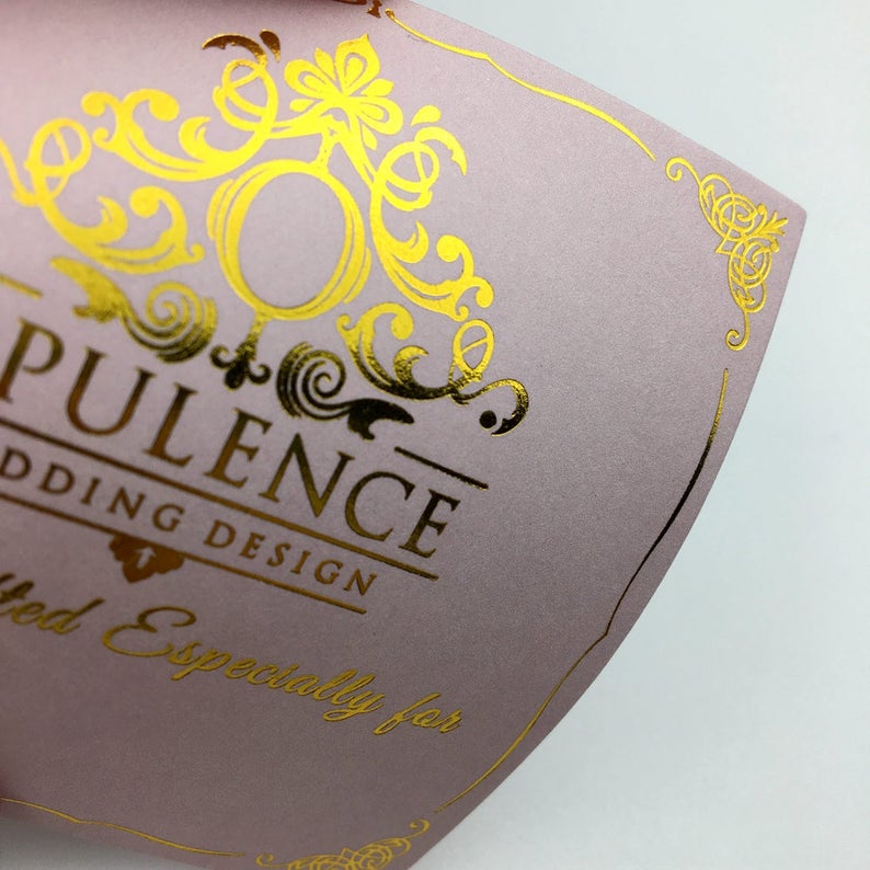 Pu&ograve; includere: Un biglietto rosa con lettere dorate in rilievo che recitano "Opulence Wedding Design" e "Creato appositamente per". Il biglietto ha un design in lamina dorata con un motivo floreale.