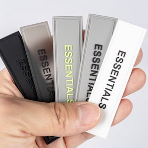 May include: Five fabric patches with the word "ESSENTIALS" printed in black on each. The patches are in different colors: black, gray, light gray, white, and a light gray with a yellow highlight.