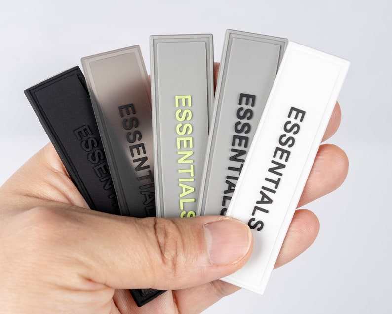 May include: Five fabric patches with the word "ESSENTIALS" printed in black on each. The patches are in different colors: black, gray, light gray, white, and a light gray with a yellow highlight.