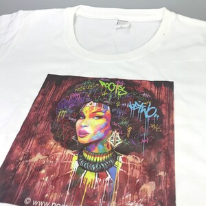 May include: White t-shirt with a colorful graphic print of a woman with an afro hairstyle. The graphic features a red, orange, yellow, green, blue, and purple color scheme with white and black accents. The background is a red and white abstract design with graffiti-style text.