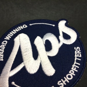 May include: A navy blue embroidered patch with white stitching. The patch is circular and features the word "Aps" in a stylised font. The text "Award Winning" and "Shopfitters" are also visible around the edge of the patch.