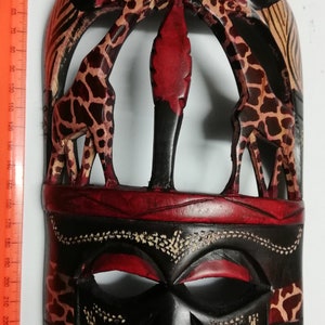 May include: A wooden mask with a black face and red and black geometric patterns. Two giraffes are carved into the top of the mask, with a red background and black and white spots.