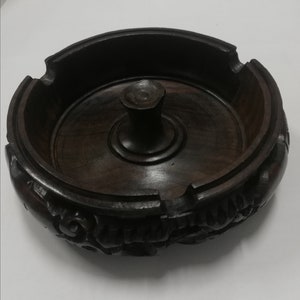 Op de afbeelding: Een donkerbruine houten asbak met ingewikkelde houtsnijwerk rond de rand. De asbak heeft een centraal, rond, verhoogd gedeelte met een kleine, ronde knop in het midden.