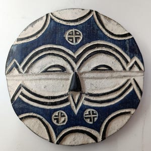 May include: A round, carved wooden mask with a face-like design. The mask features a white base with blue and black accents. The design includes geometric shapes and cross-like symbols, creating a tribal or folk art aesthetic.
