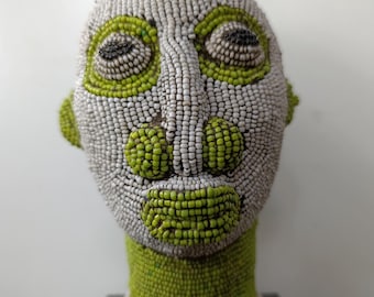 May include: A beaded sculpture of a human head. The face is primarily gray with green accents around the eyes, mouth, and ears. The neck and a small topknot are also green. The sculpture is mounted on a black base.
