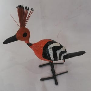 May include: A hand-crafted bird figurine with a black, white, and orange paint scheme. The bird has a long, thin beak and a spiky crest. The figurine is made of paper mache and has a wire frame.
