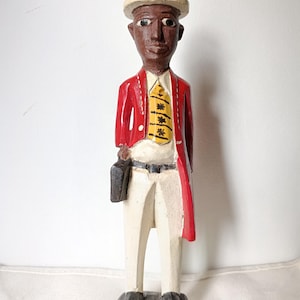 Op de afbeelding: Een gesneden houten beeldje van een man met een crèmekleurige helm, rode jas, witte broek en gele stropdas. Hij draagt een zwarte aktetas. Het beeldje heeft een donkere huidskleur en staat op een donkergrijze voet.