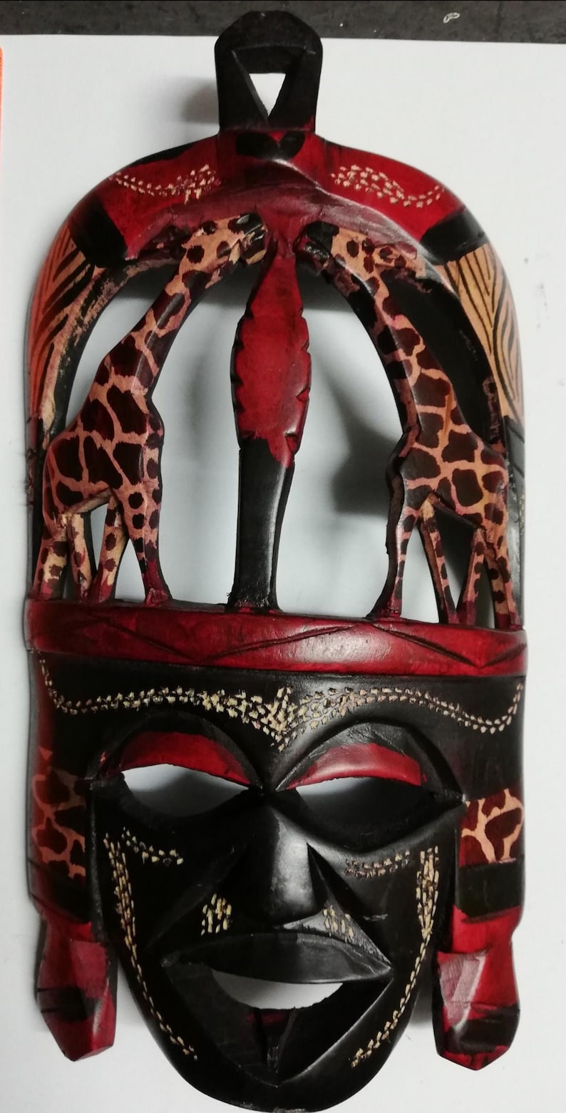May include: A wooden mask with a carved human face and two giraffes. The mask is painted in shades of red, black, and brown. The giraffes are painted in a brown and white pattern.