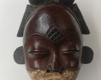 May include: A wooden mask with a dark brown face, closed eyes, and a small, pointed mouth. The mask has a geometric design on the forehead and two large, round ears. The top of the mask is decorated with a carved, black hairpiece.