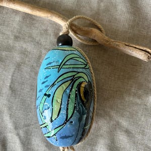 May include: An oval-shaped, hand-painted wind chime with a blue background and green and black wave design. The chime is suspended from a piece of driftwood with a natural rope. A black bead sits atop the chime.
