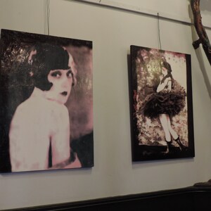 Puede incluir: Dos pinturas enmarcadas de mujeres. La pintura de la izquierda presenta a una mujer con el pelo corto y oscuro y un vestido rosa y blanco. La pintura de la derecha presenta a una mujer con un vestido negro con una falda con volantes.