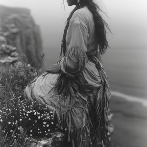 Puede incluir: Una joven vestida con ropa tradicional se sienta en un acantilado rocoso con vistas a una masa de agua. Lleva un vestido largo con flecos, un cinturón de cuentas y un tocado de plumas. Su cabello es largo y suelto, y mira hacia el agua.