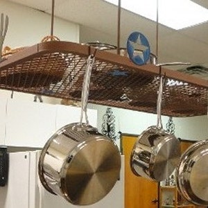 May include: A brown metal pot rack with a wire mesh shelf hangs from the ceiling. Five stainless steel pots hang from hooks on the rack. The rack has a blue star design on the front.