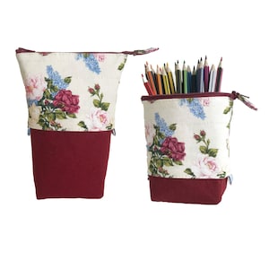 May include: Two zippered pencil cases with a floral print. The cases are white with a red floral pattern and a burgundy bottom. One case is empty and the other is filled with coloured pencils.