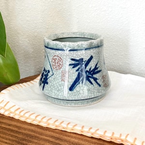 May include: A small, ceramic vase with a crackle glaze finish. The vase features blue painted bamboo and Japanese characters. The rim is slightly flared, and the vase sits on a white cloth.