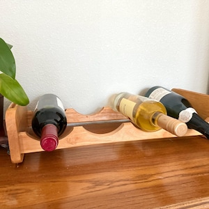 May include: A wooden wine rack holding three bottles of wine. One bottle is red wine, one is white wine, and the third is a dark bottle. The rack is made of light-colored wood and sits on a wooden surface.