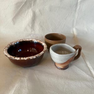 May include: Three ceramic bowls and a cup are arranged on a neutral background. The largest bowl is deep red with a textured, light-colored rim. A smaller, brown bowl and a small cup with a handle are also present.