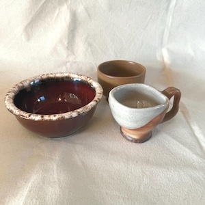 May include: Three ceramic kitchenware items: a large brown bowl with a white-speckled rim, a small brown cylindrical container, and a small cup with a handle. The cup has a white and brown color scheme.