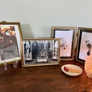 May include: A collection of framed art and keepsakes on a wooden surface. Includes a religious print, a black and white church photo, handprint art, and a footprint print. A lit candle in a textured glass holder and a small dish with matches complete the display.