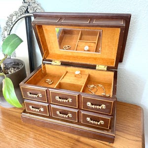 May include: A vintage wooden jewelry box with a hinged lid and multiple drawers. The box is dark brown with gold-colored hardware and a soft orange interior. The open lid reveals a mirror and compartments holding jewelry.