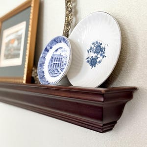 May include: A dark wood shelf displays decorative items. Two white and blue plates with floral and architectural designs are visible. A framed picture and a decorative gold-toned mirror are also on the shelf. The wall is painted a light neutral color.