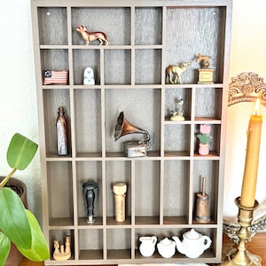 May include: A gray wooden shadow box with multiple compartments, each holding a miniature figurine. Items include a dog, an American flag, a camel, a gramophone, and a teapot. A lit candle in a brass holder is on the right.