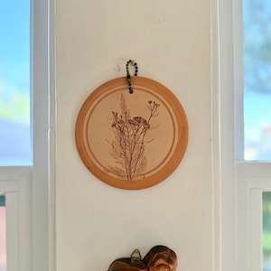 May include: A round, light brown wall hanging with a botanical design. The artwork features a detailed illustration of various plants and flowers in a naturalistic style. The hanging is suspended by a beaded chain.