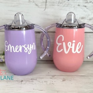 May include: Two personalized stainless steel sippy cups with clear handles and lids. The purple cup says "Emersyn" and the pink cup says "Evie".