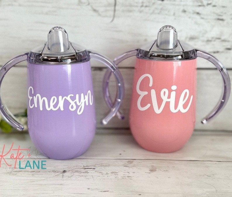 May include: Two personalized stainless steel sippy cups with clear handles and lids. The purple cup says "Emersyn" and the pink cup says "Evie".