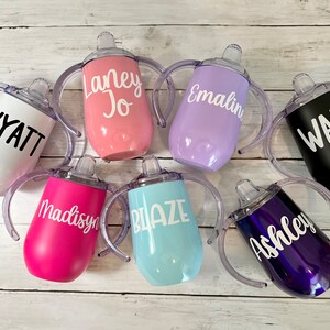 May include: Seven personalized stainless steel sippy cups with handles and clear spouts. The cups are in various colors, including white, pink, purple, black, blue, and purple. Each cup has a name written on it in white or black lettering: Wyatt, Laney Jo, Emaline, Wade, Madisyn, Blaze, and Ashley.
