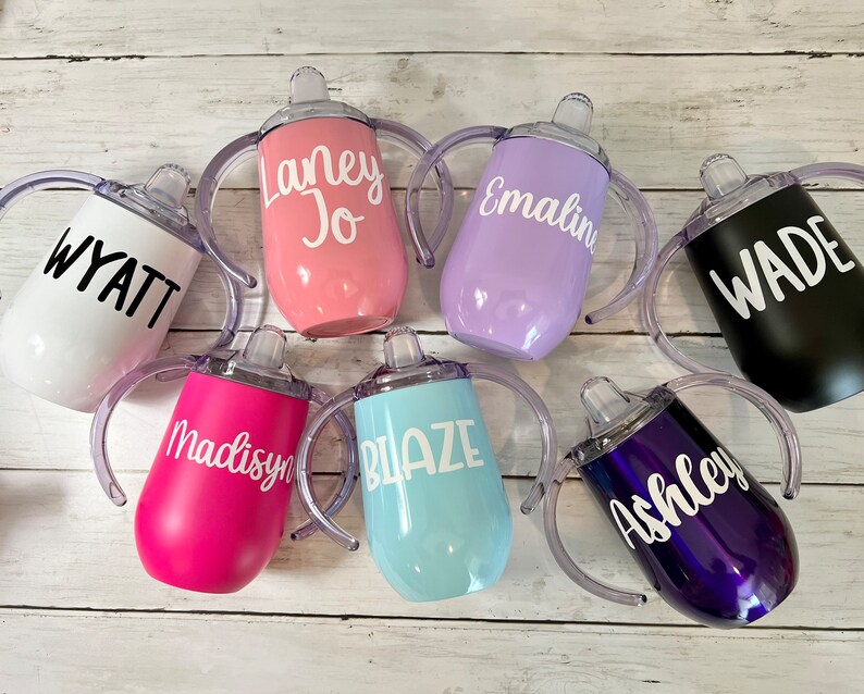 May include: Seven personalized stainless steel sippy cups with handles and clear spouts. The cups are in various colors, including white, pink, purple, black, blue, and purple. Each cup has a name written on it in white or black lettering: Wyatt, Laney Jo, Emaline, Wade, Madisyn, Blaze, and Ashley.