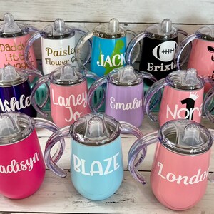 May include: A collection of 14 personalized stainless steel sippy cups with various colors and designs. The cups have two handles and a clear silicone spout. Some of the cups have names like Wyatt, Everly, Ashley, Laney, Emalin, Norah, Madisyn, Blaze, and London.