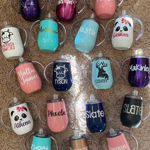 May include: A collection of 18 personalized stainless steel sippy cups with various colors, designs, and names. The cups have two handles and a clear lid.