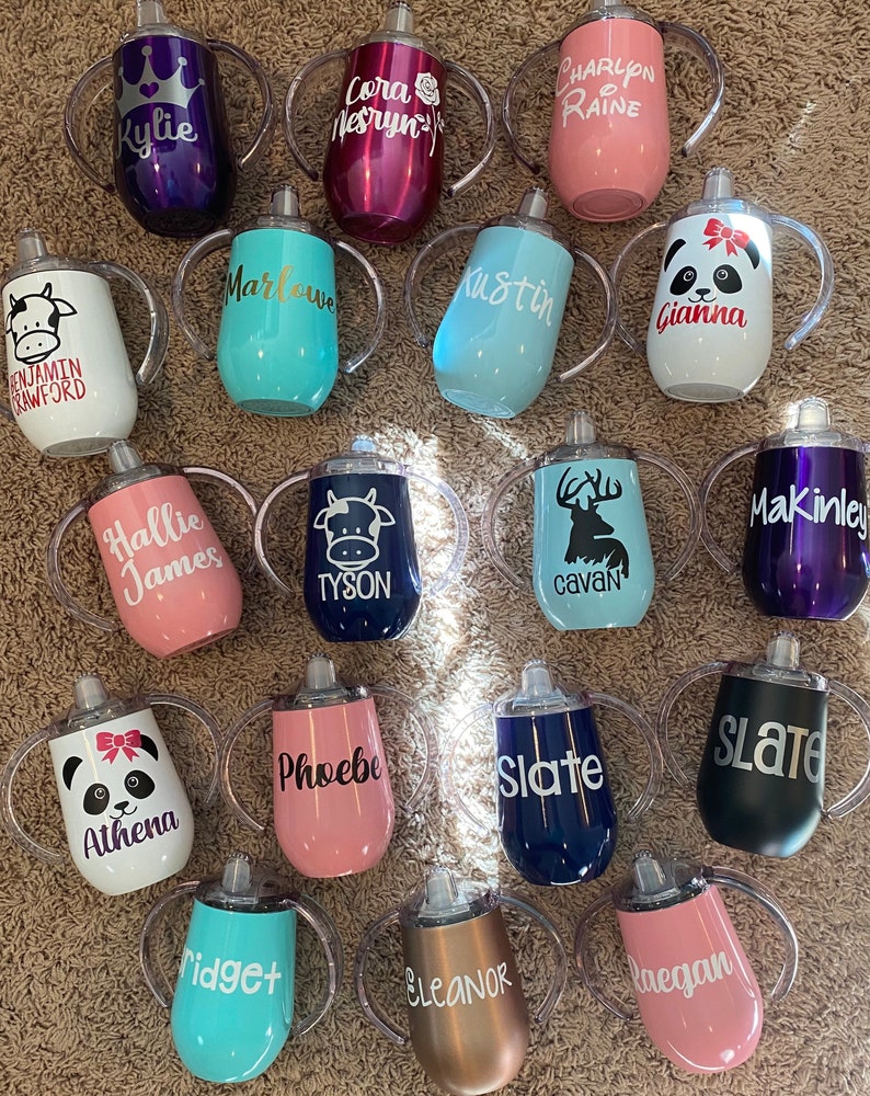 May include: A collection of 18 personalized stainless steel sippy cups with various colors, designs, and names. The cups have two handles and a clear lid.