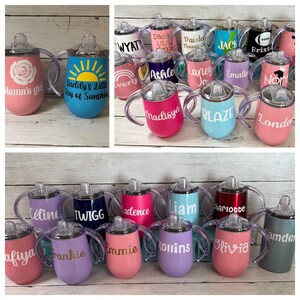 May include: A collection of 20 personalized stainless steel sippy cups with various colors and designs. The cups have two handles and a straw lid. Some of the names on the cups include: Wyatt, Daddy's Little, Paisley Flower Girl, Jack, Brixto, Mama's Girl, Daddy's Little Ray of Sunshine, Everly, Ashle, Laney Jo, Emalin, Nor, Madisyn, Blaze, London, Celine, Twigg, Kadence, Liam, Charlotte, Samde, Afiyaa, Frankie, Emmie, Kollins, Olivia.