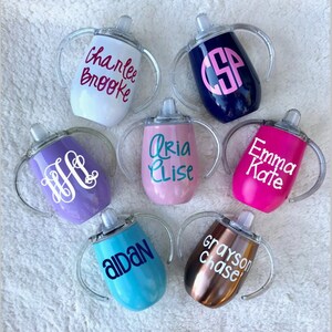 May include: Seven personalized stainless steel sippy cups with clear handles. The cups are in various colors, including white, blue, purple, pink, teal, and copper. Each cup has a different name or monogram on it, including "Charlie Brooke", "CSP", "Aria Elise", "Emma Kate", "H", "Aidan", and "Grayson Chase".