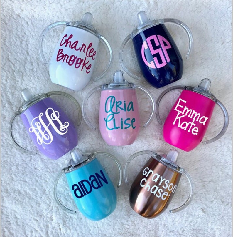 May include: Seven personalized stainless steel sippy cups with clear handles. The cups are in various colors, including white, blue, purple, pink, teal, and copper. Each cup has a different name or monogram on it, including "Charlie Brooke", "CSP", "Aria Elise", "Emma Kate", "H", "Aidan", and "Grayson Chase".