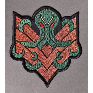 Puede incluir: Parche bordado en verde y marrón con un pulpo estilizado con ojos rojos. El pulpo está rodeado de un patrón geométrico.