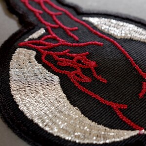 May include: Black embroidered patch with a red design of a hand with a silver crescent moon.