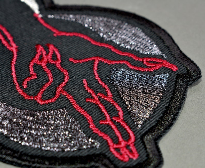 May include: Black embroidered patch with a red outline of a hand reaching out. The patch has a silver glitter design.