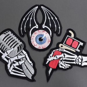 Könnte beinhalten: Drei gestickte Aufnäher auf dunklem Hintergrund. Einer zeigt eine Skeletthand, die ein Mikrofon hält, ein anderer eine Tätowierpistole und der dritte ein detailliertes Auge mit Fledermausflügeln.