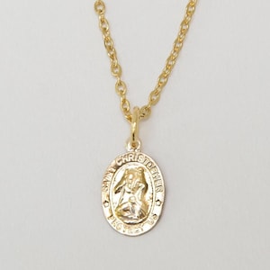 Könnte beinhalten: Eine goldfarbene Halskette mit einem Anhänger, der den Heiligen Christophorus zeigt. Der ovale Anhänger hängt an einer feinen Kette. Der Anhänger trägt die Inschrift "SAINT CHRISTOPHER PROTECT US" um das Bild herum.
