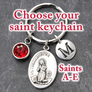 May include: Silver keychain with a red gemstone, a silver charm with the image of Saint Bernadette, and a silver charm with the letter M. Saints A-E.