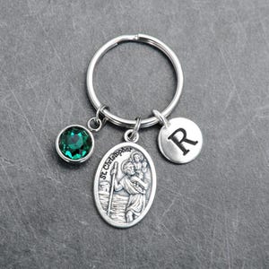 May include: A silver-toned metal keychain featuring three charms: a round emerald green crystal, an oval St. Christopher medal with an embossed figure carrying a child, and a small circular disc engraved with the capital letter R.