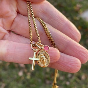 May include: Gold chain necklace with a small cross charm, a round charm with a depiction of the Virgin Mary, and a pink gemstone.