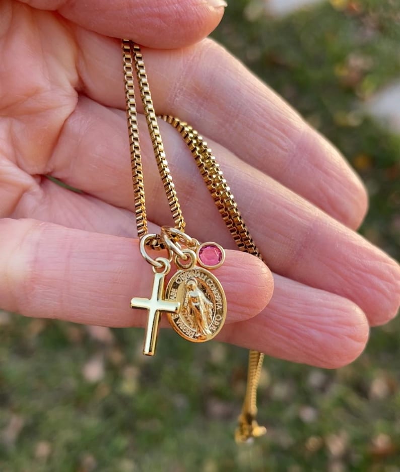 May include: Gold chain necklace with a small cross charm, a round charm with a depiction of the Virgin Mary, and a pink gemstone.