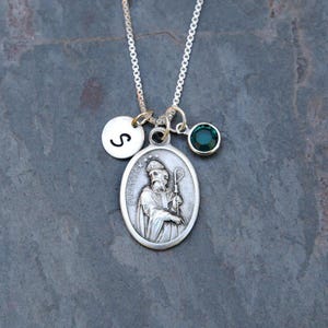 May include: A silver-toned necklace featuring an oval pendant embossed with a figure holding a staff and the text ST. PATRICK. The necklace includes a small round charm with the letter S and a circular charm with a dark green gemstone.