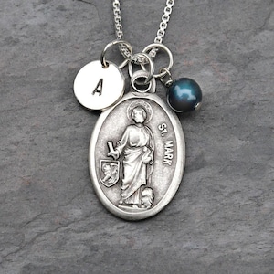May include: A silver necklace featuring a pendant with the image of St. Mark, a round charm with the letter "A", and a dark blue pearl. The pendant is oval-shaped with the words "St. Mark" on the side. The necklace is on a silver chain.