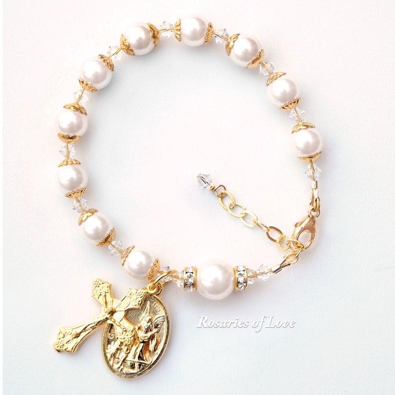 Op de afbeelding: Wit parel en kristal rozenkrans armband met een gouden engel en kruis bedel. De armband heeft een gouden sluiting en is versierd met heldere kristallen.
